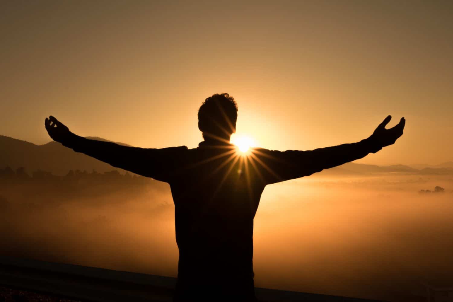 Silhouette of a person with upraised arms before the orange glow of the setting sun.