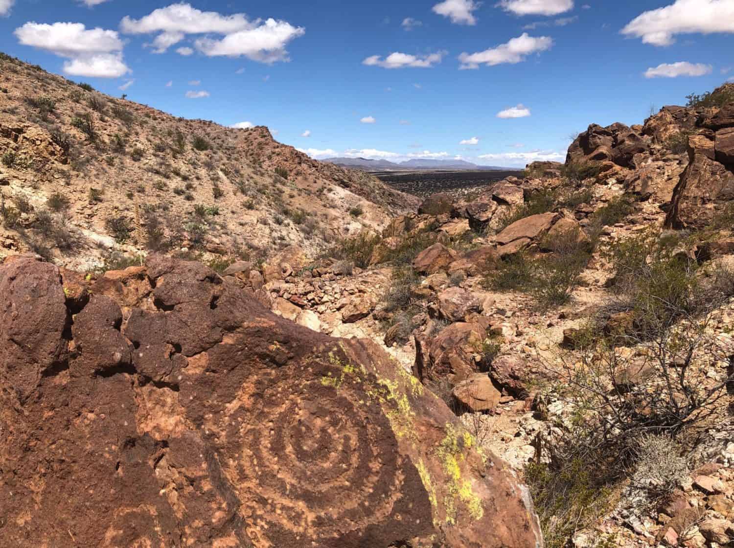 Big blue sky with white fluffy clouds, long view on mountains. Sparse vegetation on rocky hills and spiral petroglyph in the foreground.