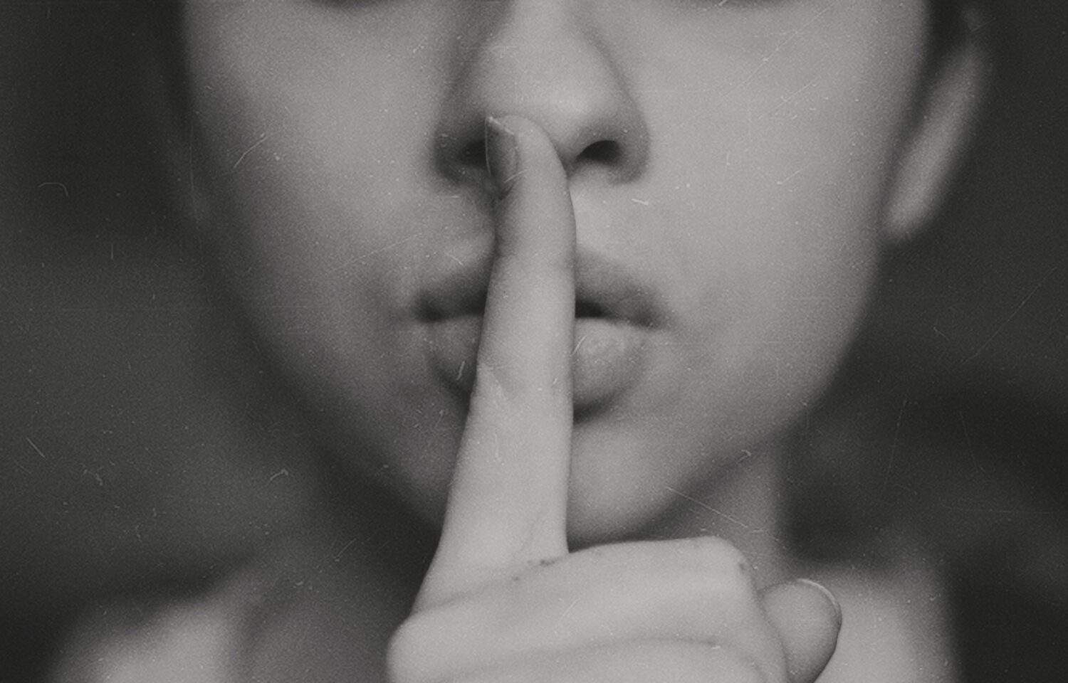 Close-up of the lower part of a woman's face with her index finger placed perpendicular to her lips, symbolizing secrets